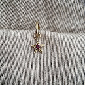 May include: A gold earring featuring a star-shaped pendant with a central red gemstone. The earring hangs against a textured, neutral-toned fabric background.
