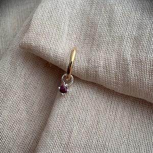 May include: A small hoop earring featuring a polished gold-tone band and a silver-tone charm. The charm consists of a circular silver loop holding a single, round, deep red gemstone set in a four-prong metal setting, creating a two-tone jewelry piece.