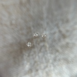 May include: Three small, square-shaped diamond stud earrings, each with a single, sparkling stone. The earrings are set in silver or white metal, and are shown against a soft, textured, light grey background.