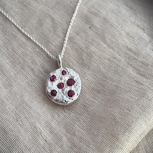 May include: A silver pendant necklace featuring a circular, textured silver disc with a rough, hammered finish. The pendant is set with five small, round, deep red rubies embedded across the surface and hangs from a delicate silver chain.