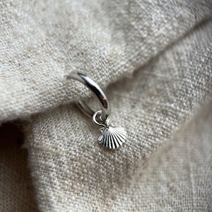 May include: A silver hoop earring with a small silver seashell charm hanging from it. The earring is on a beige fabric background.