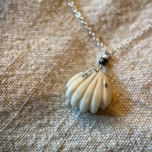 May include: A seashell pendant necklace with a silver chain. The pendant is a cream color with black speckles and a shell-like design. The necklace is on a textured, neutral-colored surface.