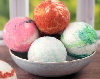 May include: Five colorful wool dryer balls in a gray bowl. The balls are pink, orange, green, and two shades of blue.