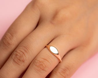 Könnte beinhalten: Ein goldfarbener Ring mit einem ovalen, weißen Mittelstein. Der Ring befindet sich an einem Finger vor einem rosa Hintergrund. Das Band des Rings ist dünn und verjüngt sich nach hinten.