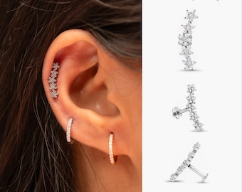 May include: A close-up of a person's ear with three different earrings. The top earring is a single row of small, clear, flower-shaped stones. The middle earring is a small, clear, hoop earring. The bottom earring is a larger, clear, hoop earring. The earrings are all set in silver.