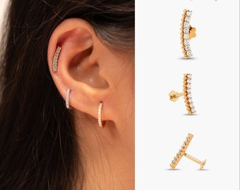 May include: Gold-toned earrings with small, sparkling stones. The image shows a curved bar earring, a small hoop earring, and a stud earring. The earrings are displayed on an ear and against a white background. The brand name "DCB Jewels" is visible.