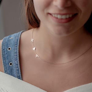 May include: A delicate silver necklace featuring the letters "R" and "A", separated by a heart-shaped charm. The necklace is worn around the neck and is a personalized piece of jewelry.