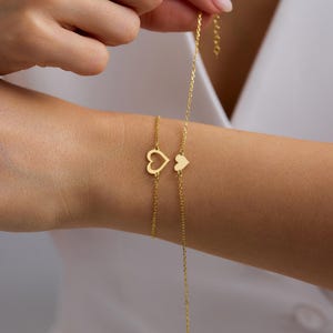 May include: Two gold-toned chain bracelets featuring heart-shaped charms. One bracelet displays an open-work heart outline, while the other features a solid, filled-in heart charm. Both delicate chains are crafted with a polished finish.