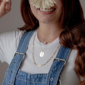 May include: A layered gold necklace set. The set includes a paperclip chain, a pendant necklace with a floral design, and a delicate pearl necklace. The necklaces are displayed on a white t-shirt and denim overalls.