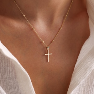 May include: A gold-colored necklace featuring a simple cross pendant. The chain is delicate, with small, evenly spaced beads. The cross hangs from a decorative bail, creating a classic and elegant look.