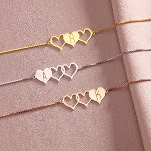 May include: Three delicate box chain bracelets in gold, silver, and rose gold tones. Each bracelet features a central design of three interconnected heart shapes, with one heart engraved with a single uppercase letter: H, A, or K respectively.