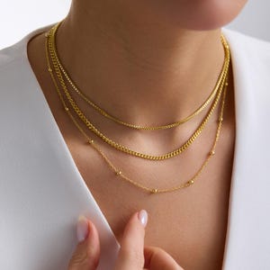 May include: A layered gold necklace set. The necklaces feature different chain styles, including a box chain, a curb chain, and a delicate chain with small ball accents. The jewelry is displayed against a white background.