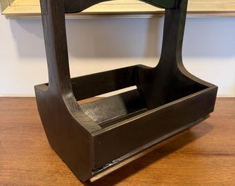May include: A dark brown wooden caddy featuring an open rectangular base with slatted sides and a high, arched handle integrated into the frame. The wood grain is visible through the dark stain, and the structure has a sturdy, simple design.