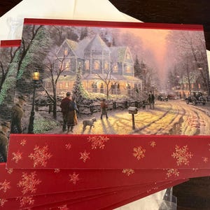 May include: Christmas greeting cards featuring a winter scene with a snow-covered house, people, and a dog. The cards have a red border with gold glitter snowflakes. An envelope is visible in the background.