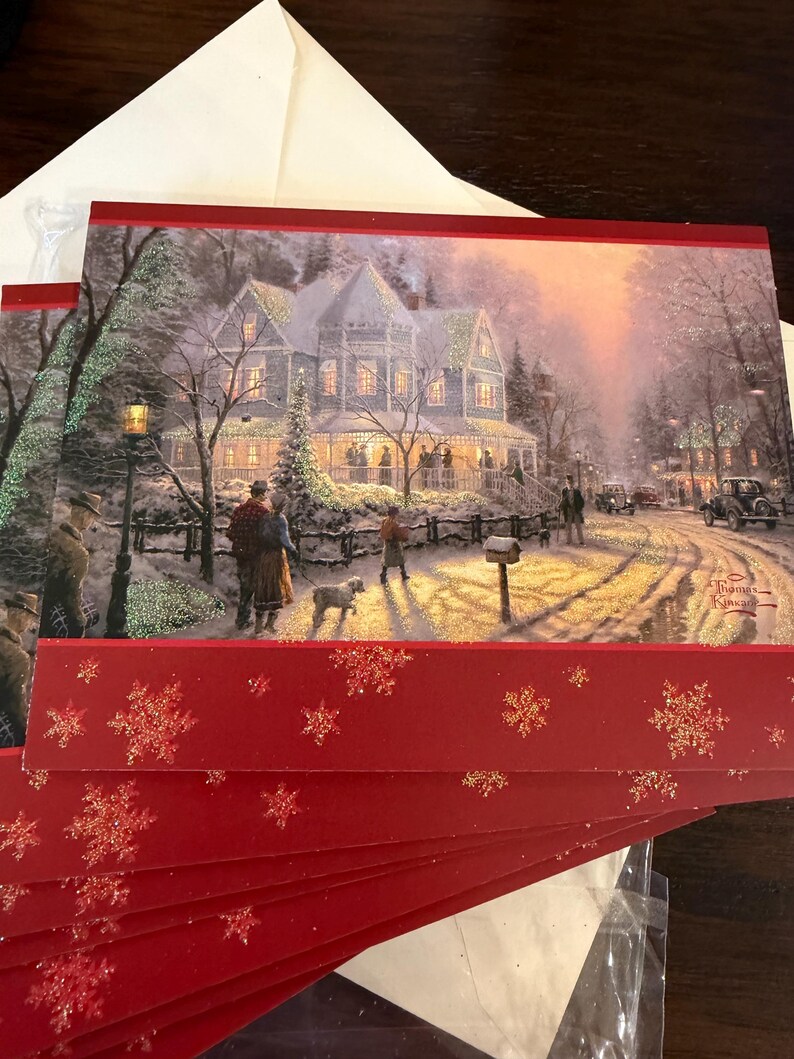May include: Christmas greeting cards featuring a winter scene with a snow-covered house, people, and a dog. The cards have a red border with gold glitter snowflakes. An envelope is visible in the background.