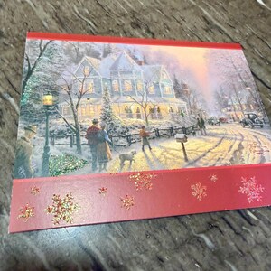 May include: A Christmas card featuring a winter scene with a snow-covered house, people, and a dog. The card has a red border with glittery snowflakes. The artwork is by Thomas Kinkade.