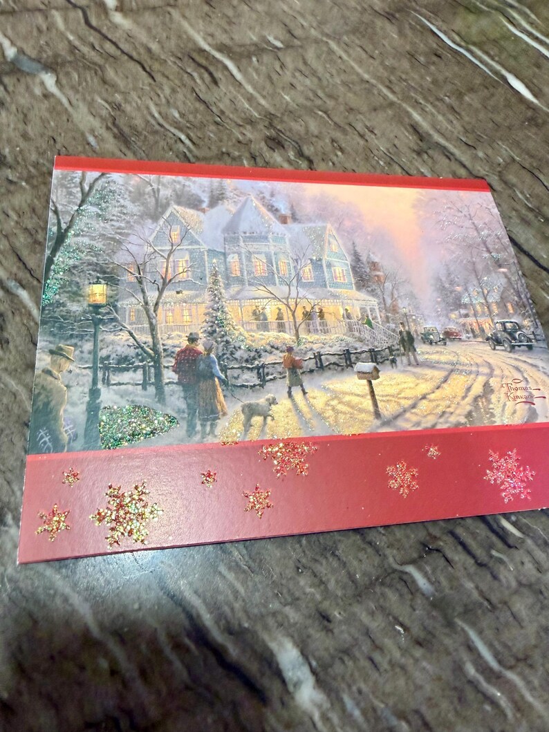 May include: A Christmas card featuring a winter scene with a snow-covered house, people, and a dog. The card has a red border with glittery snowflakes. The artwork is by Thomas Kinkade.