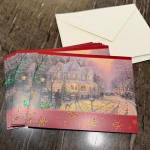 May include: A stack of Christmas cards featuring a winter scene with a house, people, and a dog. The cards have a red border with gold glitter snowflakes and come with white envelopes.