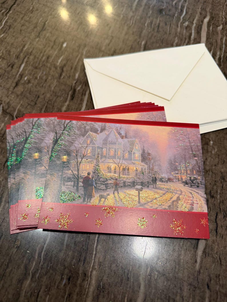 May include: A stack of Christmas cards featuring a winter scene with a house, people, and a dog. The cards have a red border with gold glitter snowflakes and come with white envelopes.