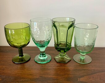 May include: Four green vintage drinking glasses with different designs. The glasses vary in shape and texture, including a textured goblet, a floral-etched glass, a faceted glass, and a textured tumbler. The glasses are arranged on a wooden surface.