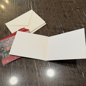 May include: An open, blank greeting card with a white interior, resting on a dark surface. A stack of cream-colored envelopes and a red and gold holiday card are also present. The card is ready for a personalized message.
