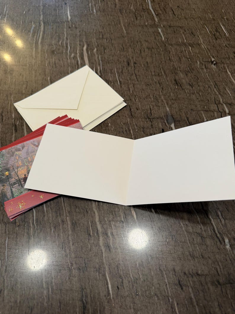 May include: An open, blank greeting card with a white interior, resting on a dark surface. A stack of cream-colored envelopes and a red and gold holiday card are also present. The card is ready for a personalized message.