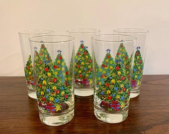 May include: Set of five clear drinking glasses. Each glass features a colorful Christmas tree design with ornaments and wrapped gifts. The glasses are arranged on a dark wooden surface.