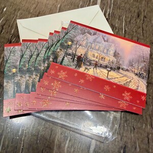May include: A set of Christmas cards with a winter scene featuring a snow-covered house and people. The cards have a red border with gold glitter snowflakes. Includes cream-colored envelopes and a clear plastic sleeve.