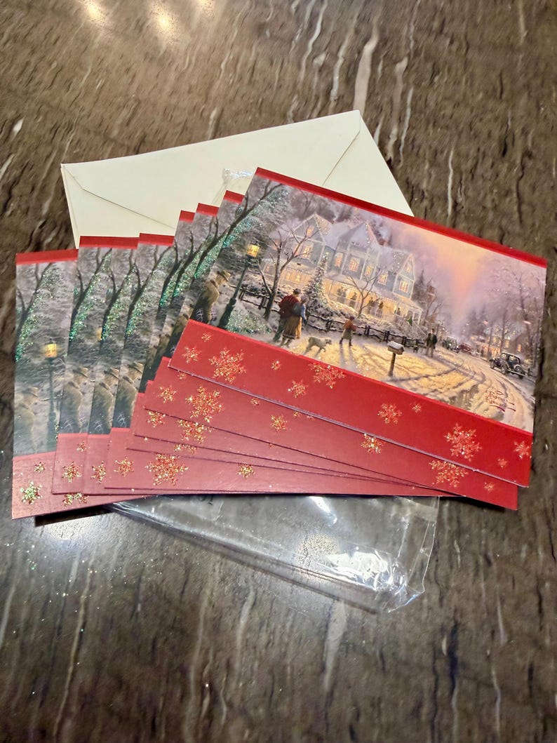 May include: A set of Christmas cards with a winter scene featuring a snow-covered house and people. The cards have a red border with gold glitter snowflakes. Includes cream-colored envelopes and a clear plastic sleeve.