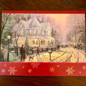 May include: A Christmas card featuring a winter scene with a snow-covered house, people, and vintage cars. The card has a red border with snowflake designs and the artist's signature.