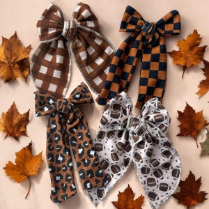 May include: Four fabric hair bows with long tails. One features a brown and white gingham pattern, one a black and orange checkerboard print, one a brown and black leopard print, and one a white fabric with a brown football pattern.