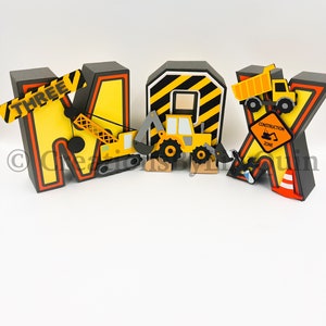 Pode incluir: Letras amarelas, pretas e laranja com tema de constru&ccedil;&atilde;o que soletram o nome "MAX" com o n&uacute;mero "THREE" na primeira letra. Cada letra tem um ve&iacute;culo de constru&ccedil;&atilde;o anexado.