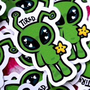 May include: A collection of green alien stickers with the word "TIRED" above their heads. The aliens have large black eyes with white heart-shaped highlights and are holding yellow star-shaped flowers. The stickers are on a pink background.