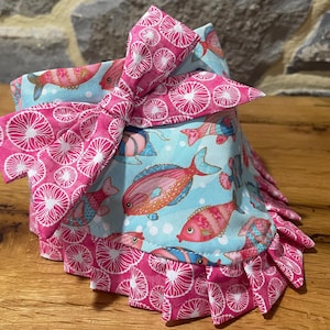 May include: A pet bandana with a pink bow and ruffled trim. The bandana features a light blue fabric with colorful fish, while the bow and trim have a pink and white circular pattern. The accessory is designed for pets.