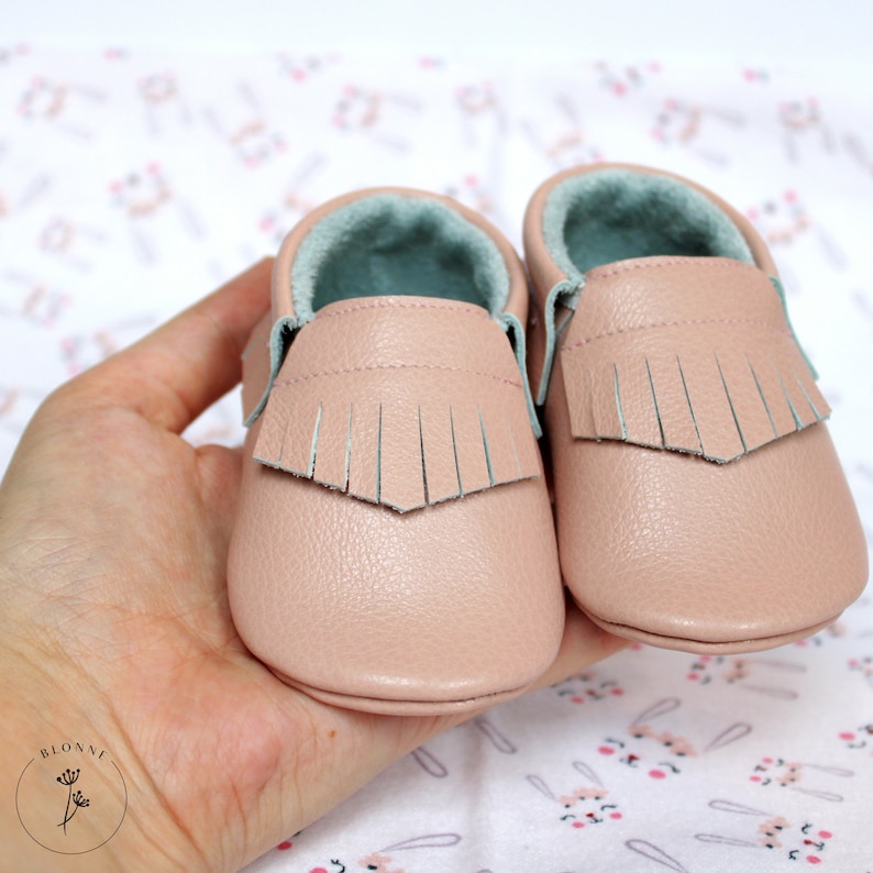 May include: A pair of pink leather baby moccasins with fringe detail. The moccasins have a light blue lining and are soft and comfortable.