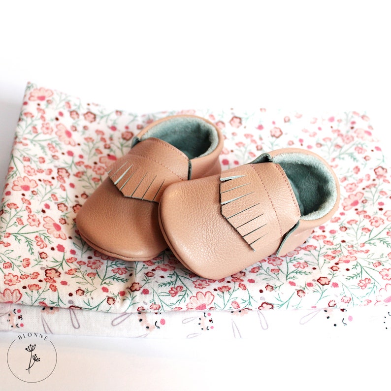 May include: A pair of light pink leather baby moccasins with fringe detail. The moccasins are on a white fabric with a floral pattern.
