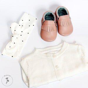 May include: A white cardigan with a button closure, a white headband with black polka dots, and a pair of pink leather baby moccasins with fringe.