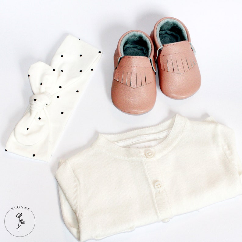 May include: A white cardigan with a button closure, a white headband with black polka dots, and a pair of pink leather baby moccasins with fringe.
