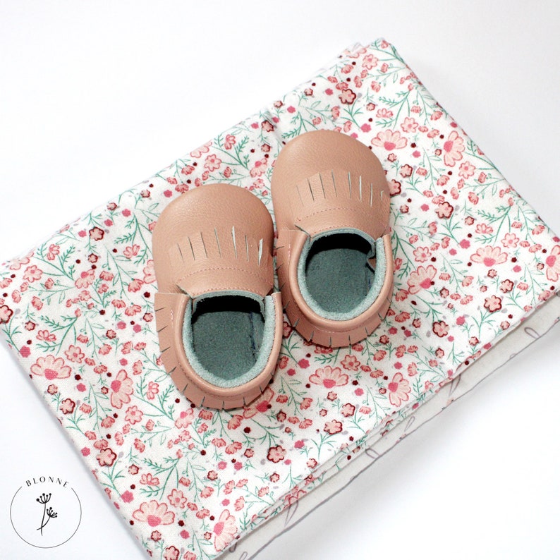 May include: A pair of light pink baby moccasins with fringe detailing on a white background with a pink floral pattern.