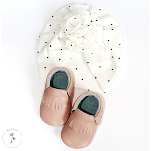 May include: A set of two pink baby moccasins with fringe detailing and a white turban with black polka dots.