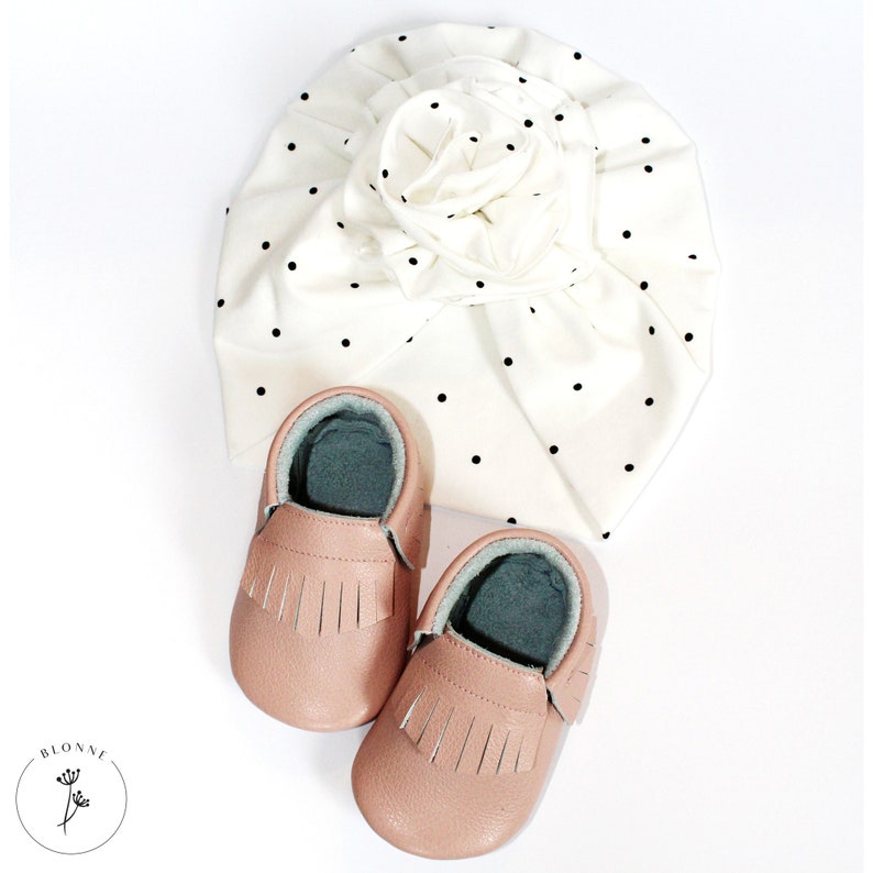May include: A set of two pink baby moccasins with fringe detailing and a white turban with black polka dots.
