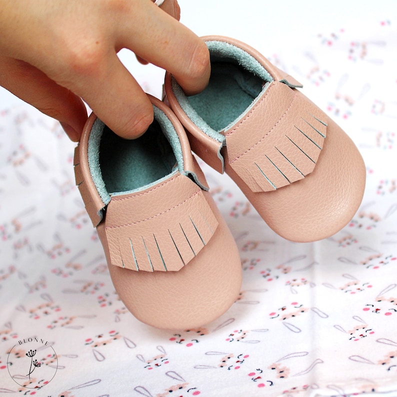 May include: A pair of pink leather baby moccasins with fringe detailing. The moccasins have a light blue lining and are shown on a white background with a bunny pattern.
