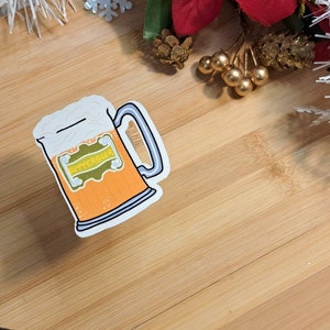 Peut inclure: Un autocollant représentant une chope de Butterbeer avec une mousse blanche et une anse bleue. La chope est orange avec une étiquette verte et dorée qui dit "Butterbeer". L'autocollant est sur une surface en bois.