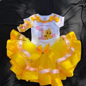 Puede incluir: Un conjunto de tut&uacute; amarillo y rosa con un body blanco. El body tiene el texto "JADE SOFIA" y un dibujo de abeja. El conjunto incluye una diadema a juego con un acento de mariposa. El tut&uacute; est&aacute; hecho de tul y cinta amarillos.