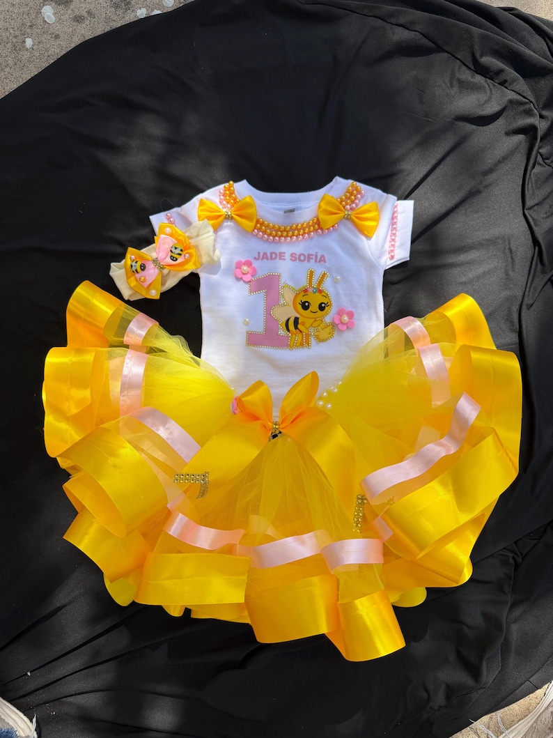 Puede incluir: Un conjunto de tut&uacute; amarillo y rosa con un body blanco. El body tiene el texto "JADE SOFIA" y un dibujo de abeja. El conjunto incluye una diadema a juego con un acento de mariposa. El tut&uacute; est&aacute; hecho de tul y cinta amarillos.