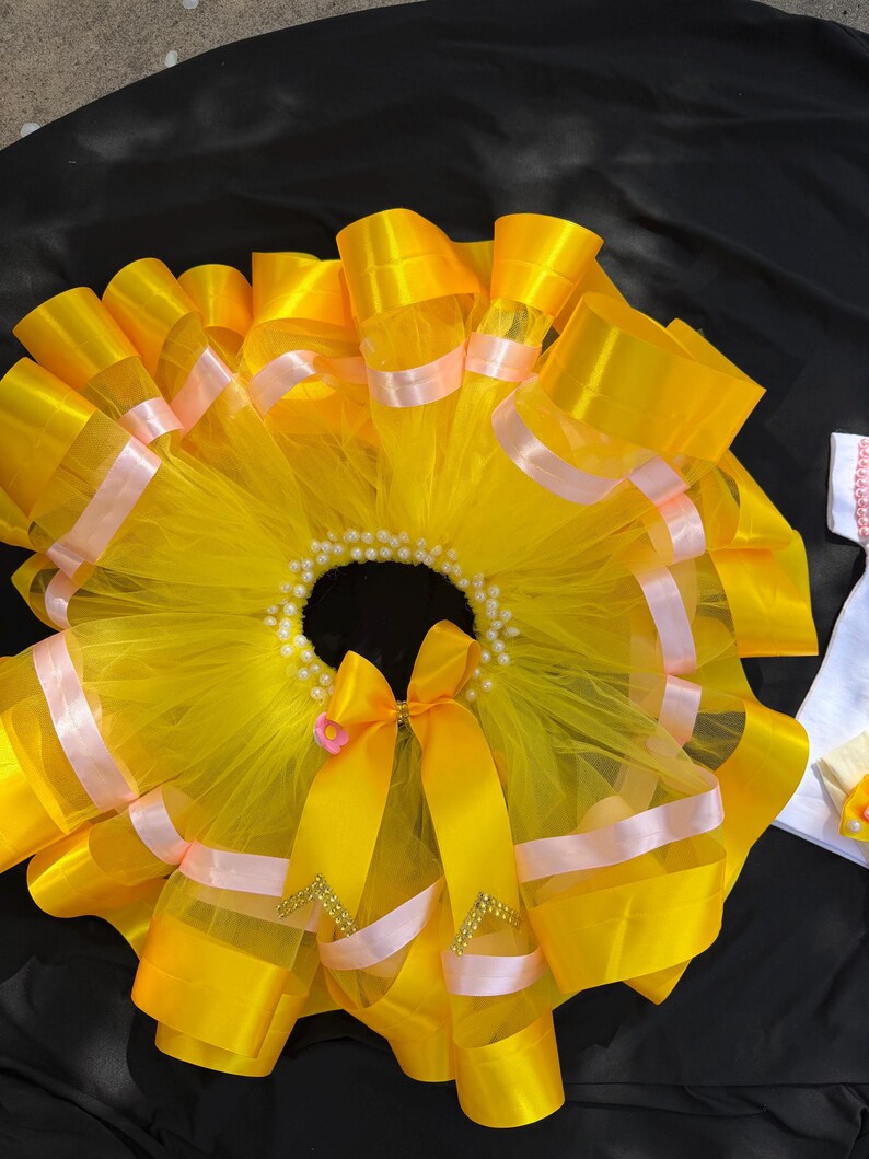 Puede incluir: Una falda tut&uacute; de color amarillo brillante con capas de tul y cinta de raso. La falda est&aacute; adornada con cinta rosa claro y una cinturilla con cuentas de perlas. Un gran lazo amarillo con una flor rosa y detalles dorados adorna la parte delantera.