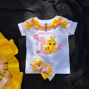 Puede incluir: Camiseta blanca con un dise&ntilde;o tem&aacute;tico de abeja, con el nombre "JADE SOF&Iacute;A" y el n&uacute;mero "1". Adornada con lazos amarillos, flores rosas y perlas. Incluye una diadema a juego con un lazo de abeja. La camiseta est&aacute; decorada con una abeja de dibujos animados.
