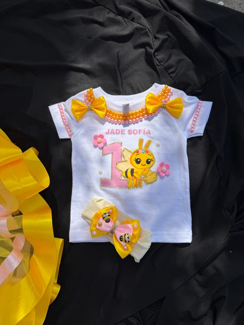 Puede incluir: Camiseta blanca con un dise&ntilde;o tem&aacute;tico de abeja, con el nombre "JADE SOF&Iacute;A" y el n&uacute;mero "1". Adornada con lazos amarillos, flores rosas y perlas. Incluye una diadema a juego con un lazo de abeja. La camiseta est&aacute; decorada con una abeja de dibujos animados.