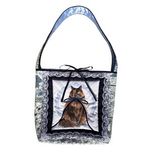 May include: A tote bag featuring a central panel with a long-haired tortoiseshell cat illustration, framed by white lace trim and dark blue fabric. The bag has a single shoulder strap and a dark blue ribbon tie closure at the top center.
