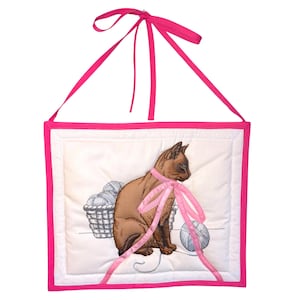 May include: A wall hanging with a brown cat illustration wearing a pink ribbon, next to a basket of yarn and a ball of yarn. The image is framed with a pink border and has a pink ribbon for hanging.